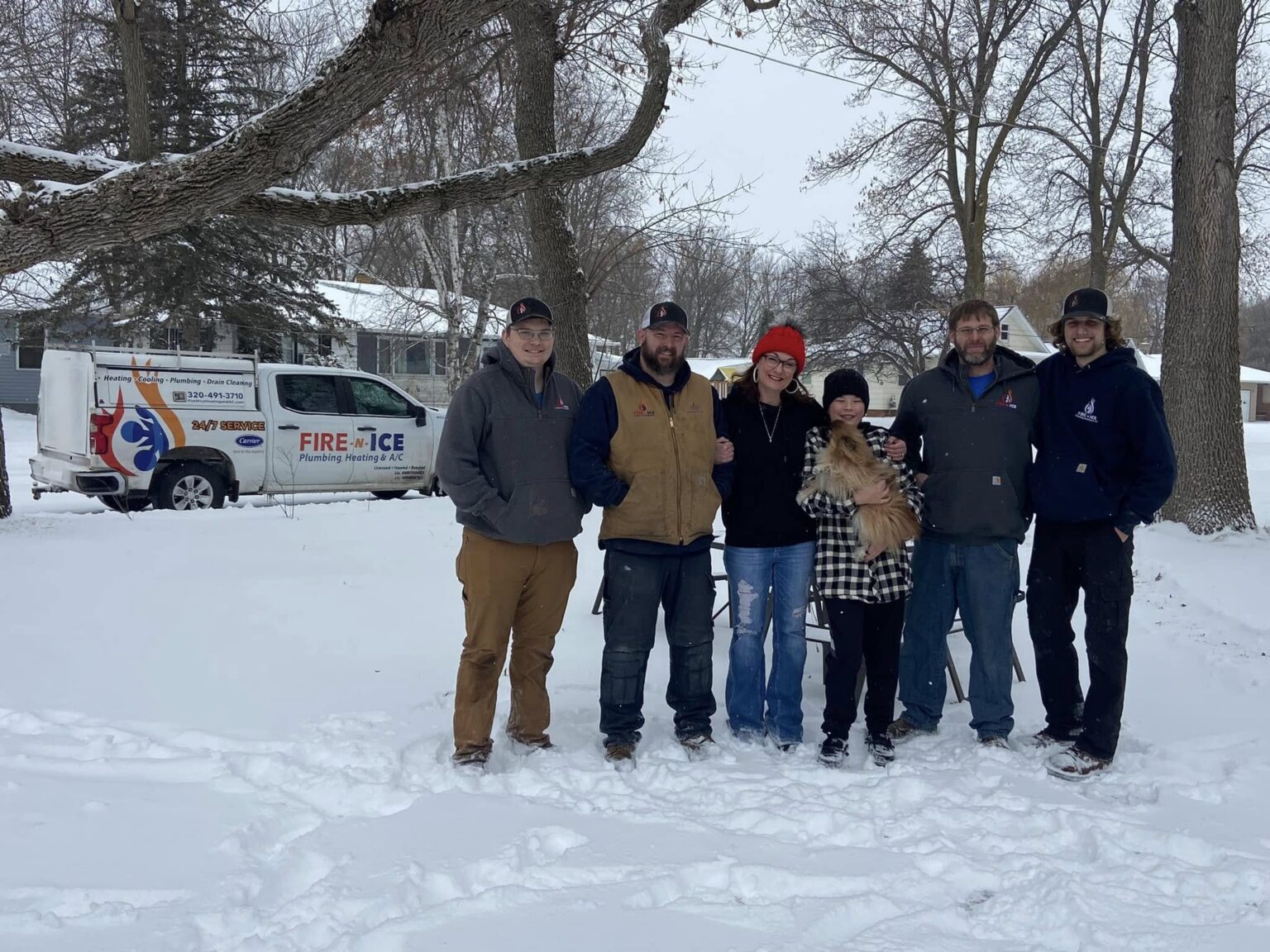 A group of six people bundled in winter jackets stand smiling in a snowy yard next to a company van, with bare trees in the background.