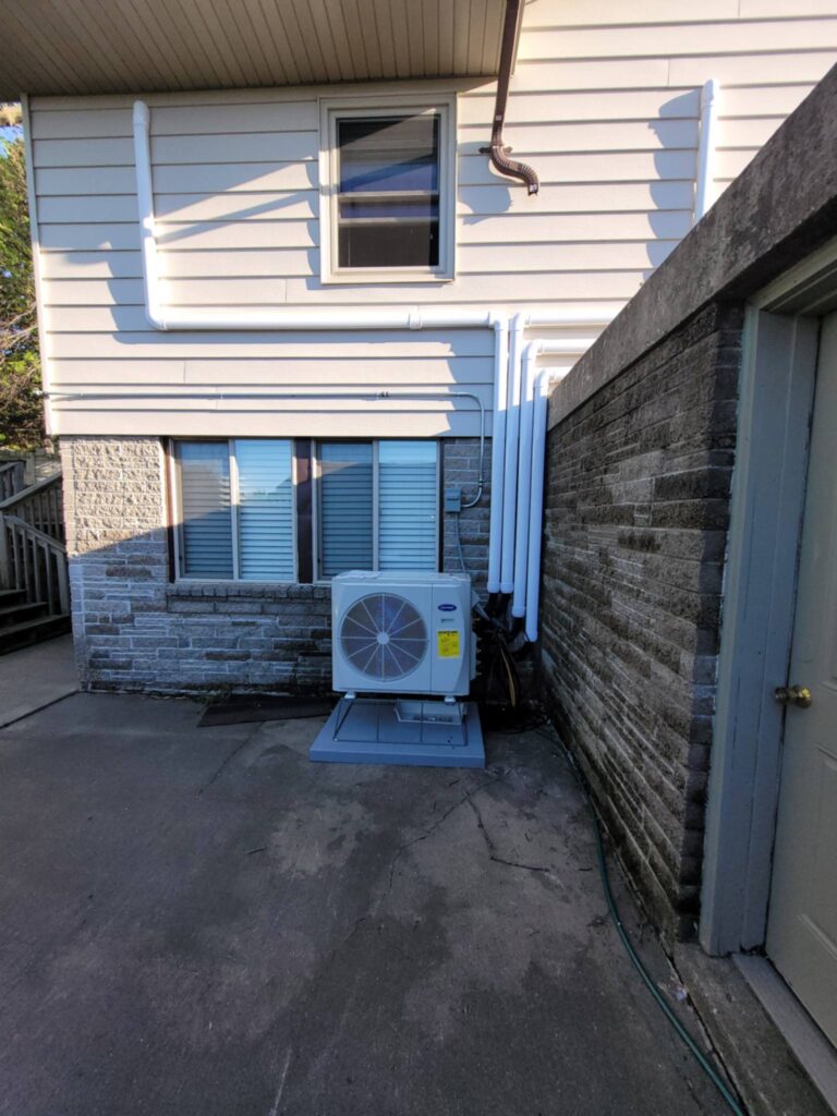 Outdoor HVAC unit installed beside a residential building, with ductwork and piping along the exterior wall.