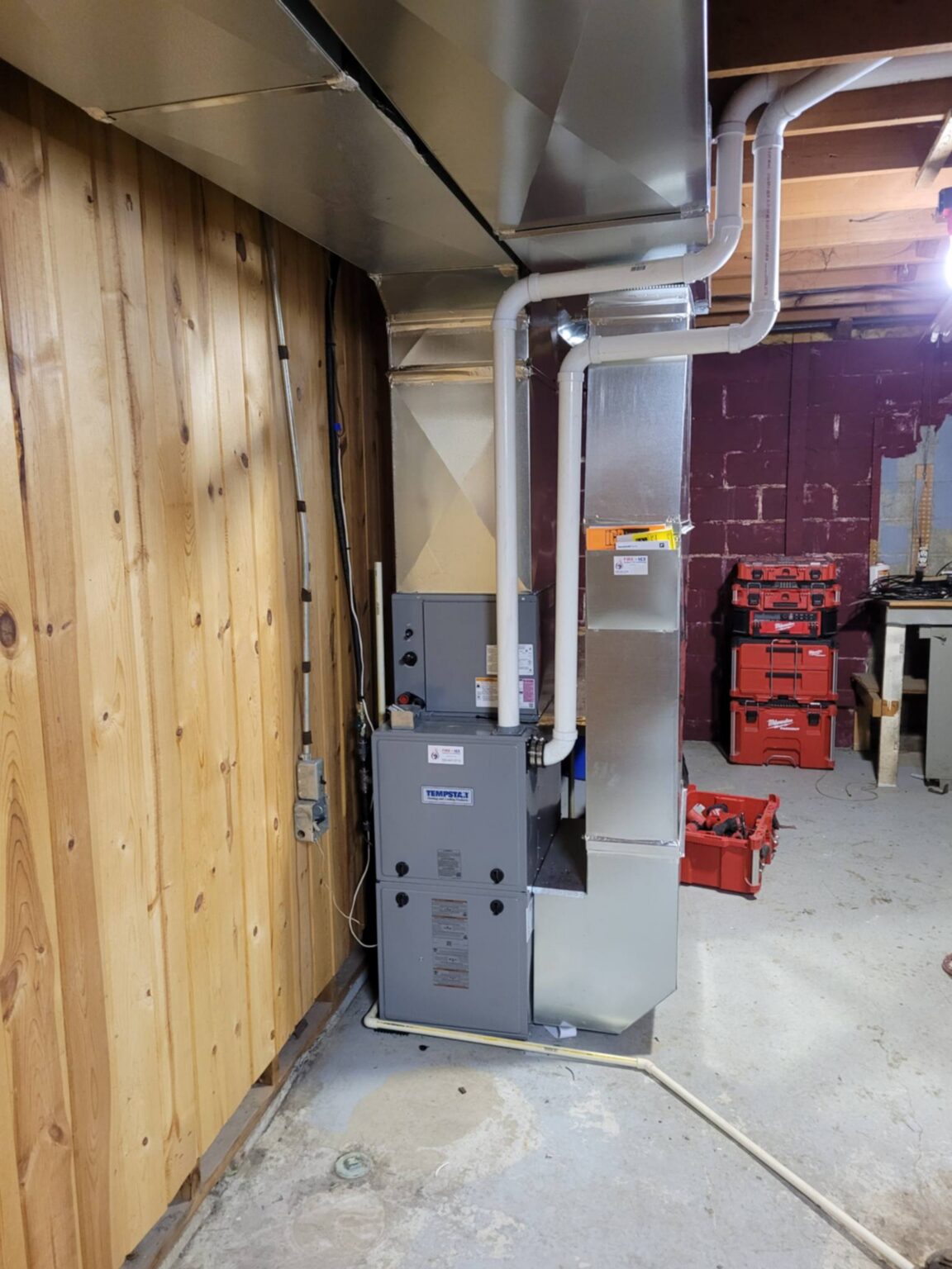 Indoor furnace and HVAC system installed against a wooden wall, with ductwork and utility equipment nearby.