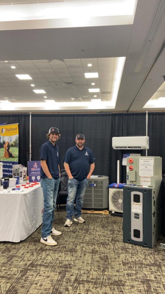 Two men in matching navy polos stand smiling at an HVAC trade show booth displaying HVAC equipemt.